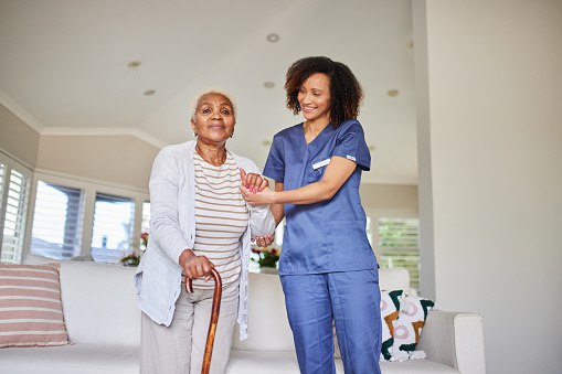 A carer from an international background, in a care home uniform, gently assists an elderly patient with a walker