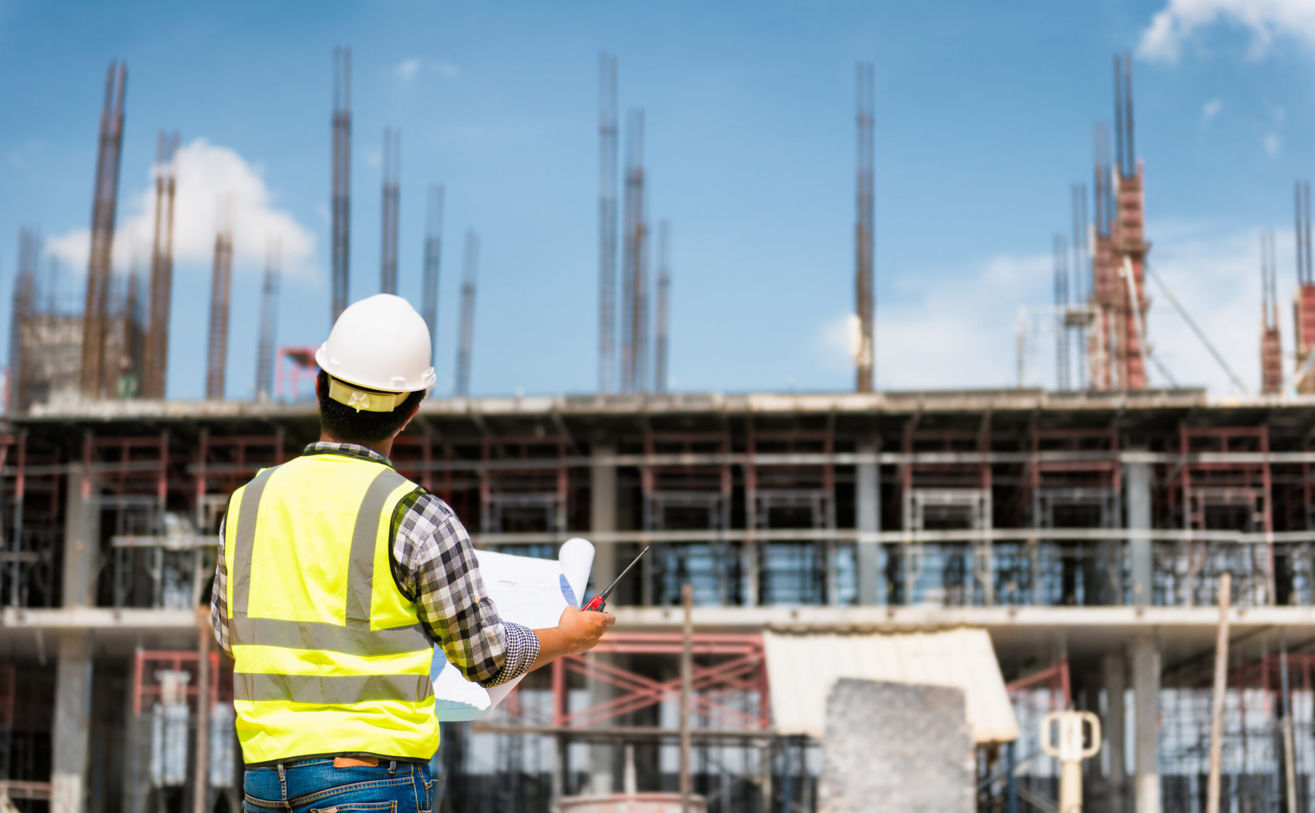 A civil engineer from an international background, in a hard hat, reviews plans on a bustling UK construction site.